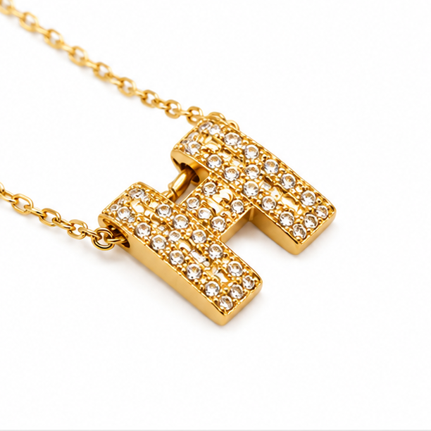 Close-up tilted shot of Zafiroon gold initial H pendant necklace encrusted with shimmering crystal rhinestones on a bright white background.