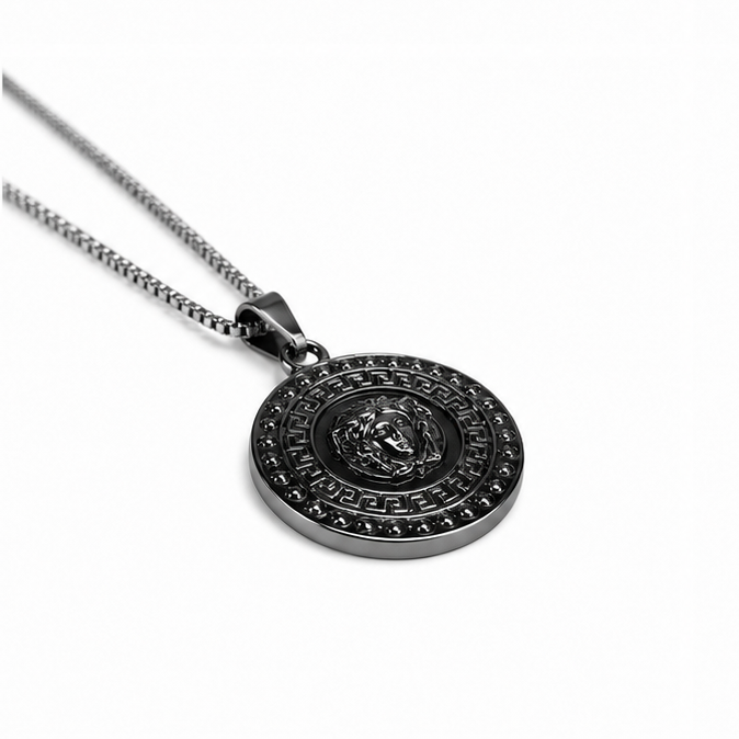 Angled perspective of an oxidized black steel round emblem necklace lying flat on a seamless white surface.